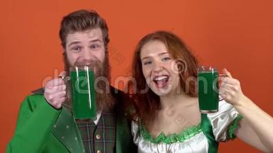 身着盛装庆祝圣帕特里克节的年轻一对夫妇`橙色的墙上隔离了一天，为绿色的啤酒干杯